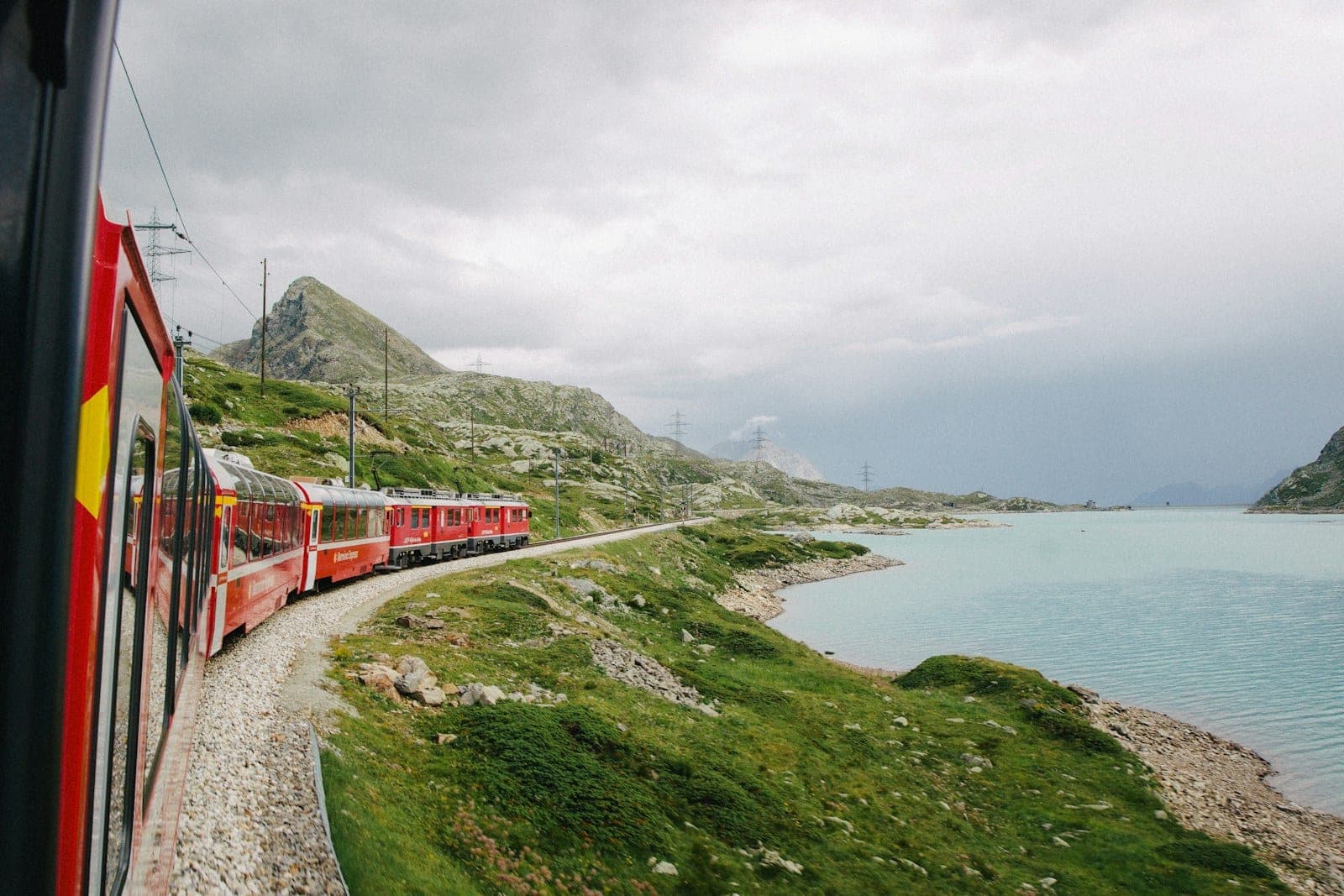A red train curving along the shore of a pale glacial lake, seen from a window