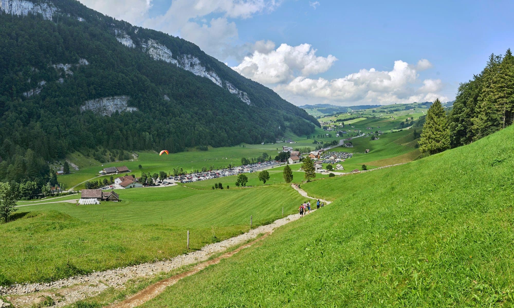 Walkers on a meadow path descending towards a village, spruce and green slopes around them